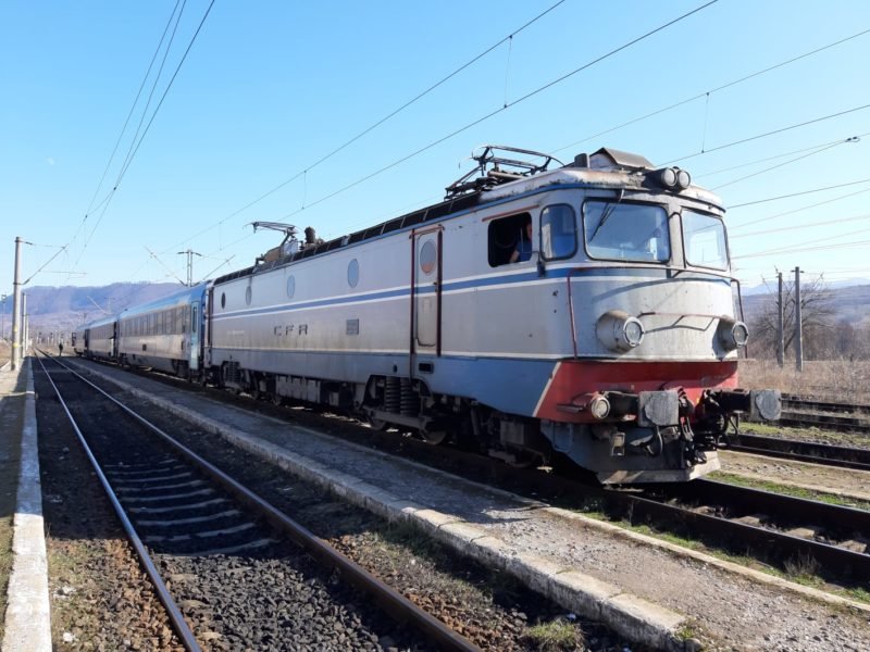 O locomotivă electrică CFR care remorcă un tren de călători într-o stație de cale ferată, pe fundal de dealuri, într-o zi senină.