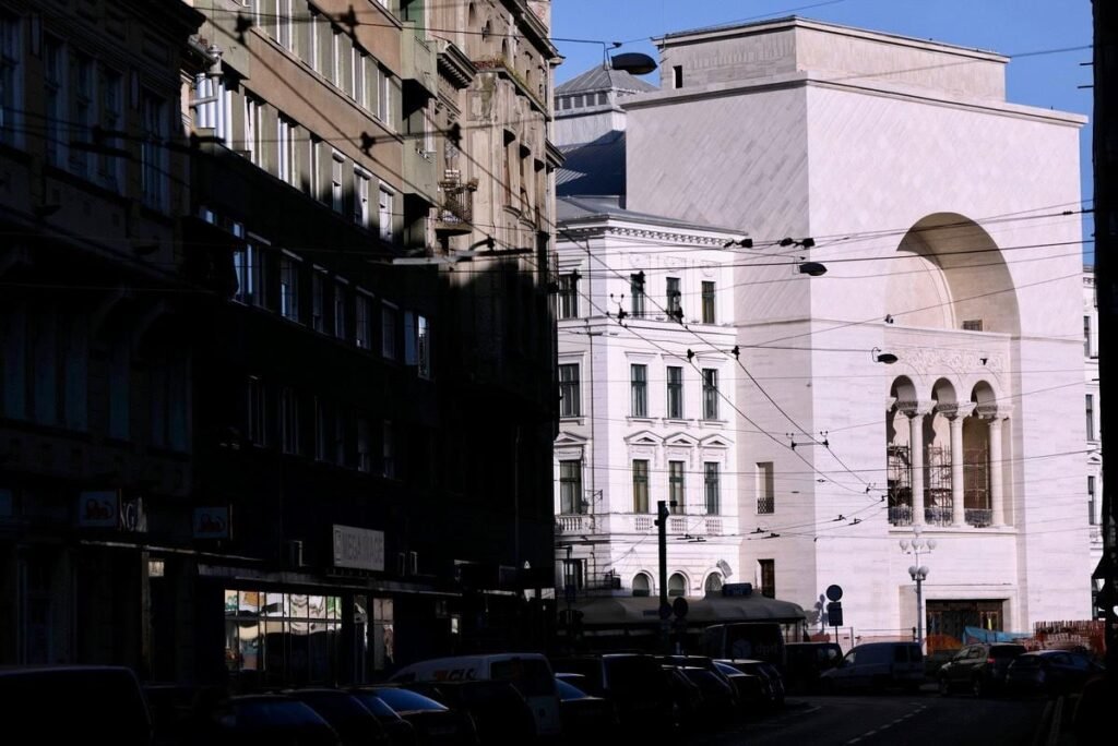 O clădire albă monumentală cu un arc mare și o logie cu coloane, într-o stradă urbană umbrită, cu mașini parcate și cabluri aeriene; seamănă cu clădirea Teatrul