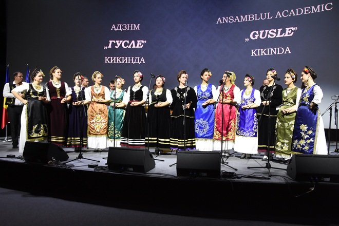 Un ansamblu folcloric feminin în costume tradiționale sârbești, pe scenă, în fața unui ecran cu textul „Ansamblul Academic Gusle Kikinda”, cu steaguri în fundal