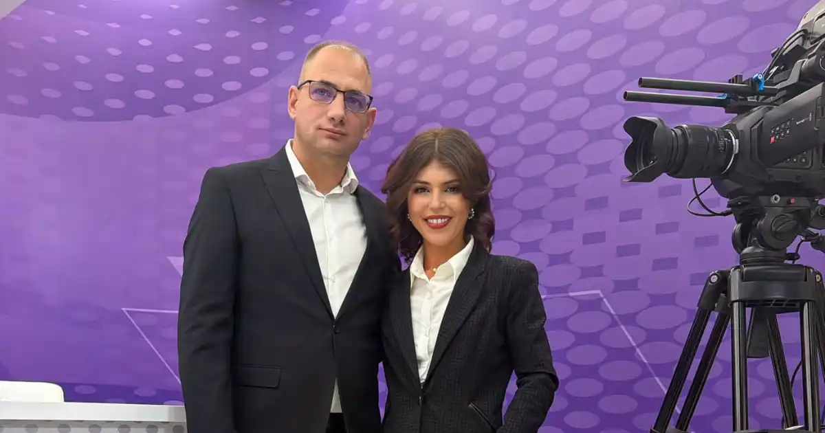Un bărbat în costum și ochelari și o femeie în sacou negru pozează într-un platou de televiziune cu fundal mov și o cameră profesională în dreapta.